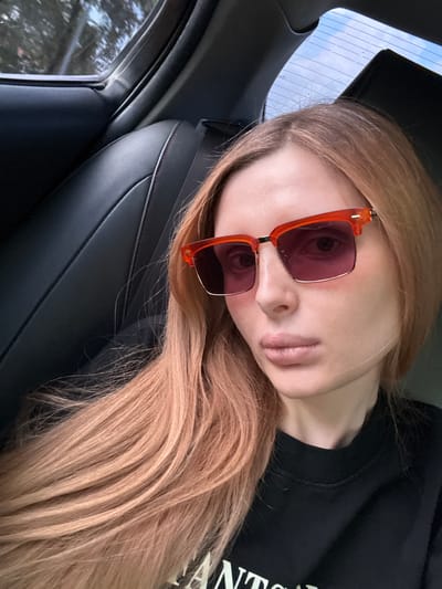 The image shows a young Caucasian woman with long, light brown hair and fair skin. She is wearing large, orange-rimmed sunglasses with pink lenses and a black t-shirt. The woman is lying down, with her head tilted and resting on her shoulder, looking towards the camera. Her lips are slightly parted.
She appears to be inside a car, as indicated by the black leather car seat and the interior of the car door visible in the foreground. Through the car window, a cloudy sky and some trees can be seen, suggesting it is daytime. The lighting is soft, possibly indicating overcast weather or late afternoon.
The overall mood of the image is casual and relaxed, possibly suggesting the woman is on a car journey. There is no visible text in the image. The location context provided is Novaya Adygeya, Russia.