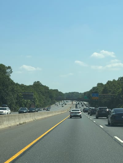 A vertical, eye-level, outdoor, daytime, full shot captures a highway scene in Glenarden, Maryland, United States of America. The sky is a bright, clear blue, with a few wispy white clouds scattered across it. The highway itself is asphalt and has a prominent yellow line down the center of the lane. Several cars are on the highway, driving in the same direction. The cars closest to the camera include a white SUV on the left, a black SUV in the center, and a dark-colored sedan on the right. Further down the highway, more cars are visible, appearing as smaller shapes. Trees line both sides of the highway, forming a dense green wall. In the distance, overpasses and highway signs are visible, indicating the complex nature of the road system. The overall impression is one of a typical, busy highway commute on a sunny day.