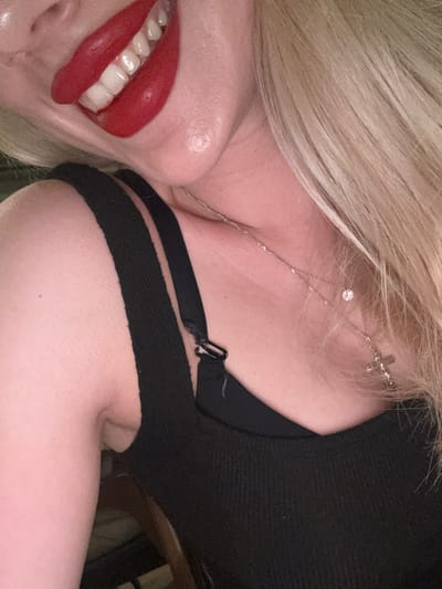 A close-up, angled image features a person with fair skin and straight blonde hair. The subject's mouth is open, revealing white teeth and the tip of their tongue, coated with a glossy, vibrant red lipstick. Blonde hair cascades over the right shoulder and upper chest. The individual is wearing a black ribbed tank top. A black bra strap with a plastic adjuster buckle is visible beneath the tank top strap on the right shoulder. A thin, silver-colored necklace with a small clear faceted stone and a small silver-colored cross pendant hangs around the neck. The skin on the arm and shoulder shows minor texture and a small dark freckle. The background is indistinct and dark, providing no specific environmental details.