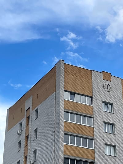 This media file is an upward-facing shot of a modern building exterior against a blue sky with scattered white clouds. The building is constructed with alternating bands of light gray and tan brickwork. The gray brick forms the primary facade and is laid in a stretcher bond pattern. The tan brick appears in horizontal bands and also in diagonal sections between rows of windows.
The windows are rectangular and recessed, with white frames. Some windows are open, showing interior blinds. On the upper section of the building, there is a circular decorative element with a horizontal line and a vertical line inside, resembling a clock face without numbers or hands. There is also a square tan brick section just to the left of this circular detail.
The building is tall, suggesting it is an office building or apartment complex. The lighting indicates it is daytime, with the sun likely positioned above and to the side of the building, casting subtle shadows. The weather appears clear and pleasant. The scene is static, with no activity or people visible. The location context is Klintsy, Russia. No text is visible in the media file.