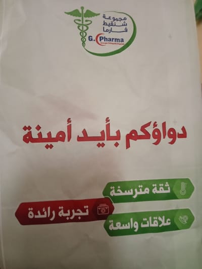 The image displays a white surface, likely a bag or a promotional flyer, featuring text and logos, predominantly in Arabic and some English.
At the top, there is a prominent logo for a pharmaceutical entity. This logo consists of a green outline forming a partial ellipse or circle. Inside this green shape, on the left, is a green caduceus symbol (a staff entwined by two snakes with wings, commonly associated with medicine). To the right of the caduceus, in Arabic script, are the words "مجموعة شنقيط فارما" (Majmouat Chinguitt Pharma), which translates to "Chinguitt Pharma Group." Below this Arabic text, in English, it says "G. Pharma" in large red letters, and directly underneath that, in smaller grey text, "Groupe Chinguitti Pharma."
Below the logo, centered on the white surface, is a large, bold phrase in red Arabic script: "دواؤكم بأيد أمينة" (Dawao'ukum bi'aīdin amīnah). This translates to "Your medicine in trustworthy hands."
Towards the bottom of the image, there are three distinct horizontal banners with rounded ends, each containing Arabic text and an icon.
1. The top-right banner is green and contains the Arabic text "ثقة مترسخة" (Thiqah mutarassikhah), meaning "Established trust," accompanied by a white thermometer icon.
2. The middle-left banner is red and contains the Arabic text "تجربة رائدة" (Tajribah rā'idah), meaning "Pioneering experience," accompanied by a white icon that appears to be a camera or possibly a briefcase/satchel.
3. The bottom-right banner is green and contains the Arabic text "علاقات واسعة" (Alaqat wasi'ah), meaning "Extensive relationships," accompanied by a white icon resembling a handshake or a conversation bubble.
The overall design suggests branding for a pharmaceutical company or pharmacy, aiming to convey professionalism, trust, experience, and good customer relations. The surface appears slightly creased, consistent with a soft material like paper or plastic for a bag.
