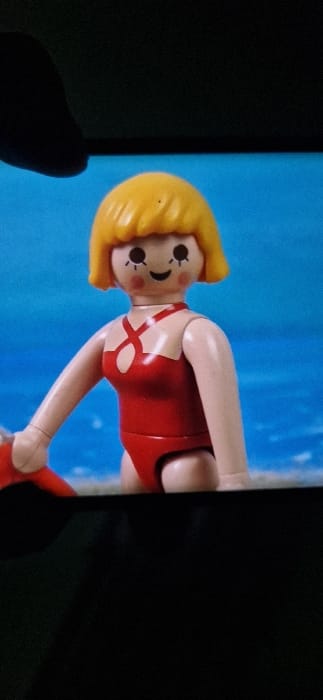 A photograph shows a Playmobil figurine of a woman with blonde hair and a red swimsuit, displayed on a smartphone screen. The background is a blurred blue, possibly representing water. Part of the phone's black bezel is visible, along with a hand holding the device. The image is partially obscured by shadow, particularly on the right side.
