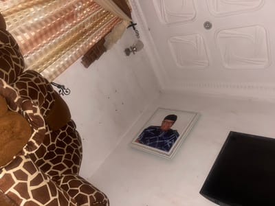 The image displays an indoor scene, possibly a living room, in Kporva, Nigeria. The room features white walls and a decorative white ceiling with square recessed panels and a central light fixture. A framed portrait of a man wearing a dark patterned outfit and a black cap hangs on the wall. To the left, a large, plush giraffe-themed stuffed animal or furniture piece is visible, partially obscuring the wall and curtains. The curtains, visible at the top left, are sheer with a horizontal striped pattern in muted tones of peach and beige. A wall-mounted light fixture with a metallic shade is also present near the curtains. The overall impression is of a residential interior with personal decorative elements.