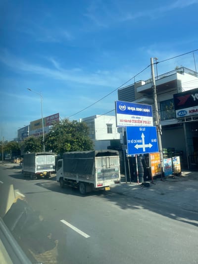 This is a photograph taken from inside a vehicle, looking out onto a street in Diên Lạc, Vietnam. The scene is a roadside with buildings and traffic.

The primary subjects are several trucks parked or moving along the road. One truck in the foreground is white with a black tarp covering its cargo. Another truck further down the road also appears to be white with a similar tarp. There are also various signs visible along the roadside. One prominent blue sign indicates directions to Dalat, Ho Chi Minh City, and Hanoi, with arrows showing the respective directions. Another large blue and white sign advertises "NHỰA BÌNH MINH" (Binh Minh Plastic) and "THIÊN PHÁT" (Thien Phat), suggesting it is a distributor or store for plastic products. Other signs, some yellow and red, are also visible, including one that mentions "SAO LIMOUSINE" and "NHA TRANG - TP. HCM," indicating a bus or transport service.

The setting appears to be a commercial area with buildings lining the street. One building has the word "YO" visible at the top, and another sign with "KH" and "KIM" is present, possibly indicating businesses. The sky is clear and blue, suggesting a sunny day. The time of day appears to be daytime, likely morning or afternoon, given the bright sunlight casting shadows on the road. The overall impression is a typical street scene in a Vietnamese town or city, with a focus on commerce and transportation. There are no people clearly visible in the frame.