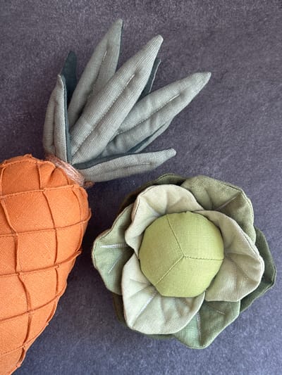 This is a high-angle, close-up, product photograph of two stuffed fabric vegetables placed on a dark gray, textured surface. On the left is an orange stuffed carrot with a grid pattern created by stitching and a burlap-like twine wrapped around its top where green fabric leaves emerge. To the right of the carrot is a stuffed green cabbage, made of layered green fabric leaves that curl outwards from a lighter green core. The background is a plain, dark, textured surface. There is no indication of the city of Chornomorske, Ukraine.