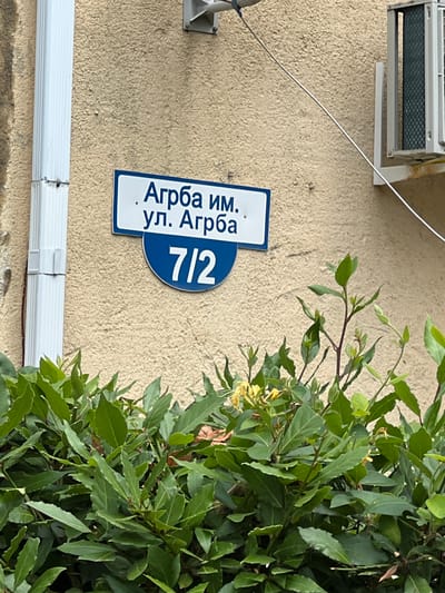 The image shows a street sign for "Агрба им. ул. Агрба 7/2" in Pitsunda, Abkhazia. The sign is mounted on a textured beige wall. Below the sign, lush green foliage obscures the lower portion of the wall and the ground. To the left of the sign, a white downspout is visible. To the right, an air conditioning unit is mounted on the wall, with a thin metal wire extending diagonally across the image from near the top left to the top right. The scene appears to be outdoors during daylight.