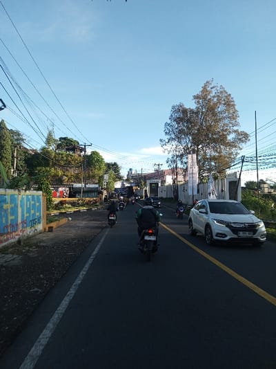 The image displays a daytime street scene in the city of Khouathineung, Laos, under a clear blue sky. The asphalt road features a solid white line on its left edge and a solid yellow line separating the directions of traffic.
Multiple vehicles are present. In the immediate foreground, a dark-colored motorcycle is centrally positioned, ridden by an individual wearing a dark jacket over a green vest and a dark backpack. The motorcycle's rear license plate reads "3594 NH." Several other motorcycles are visible ahead, traveling in the same direction. On the right side of the road, a white Honda HR-V SUV, bearing license plate "B 996 GRS," is observed traveling in the opposite direction, followed by a dark-colored sedan.
To the left of the road, a concrete barrier or wall is extensively defaced with blue and red graffiti. Behind this barrier, an elevated, densely vegetated embankment rises, featuring numerous trees and shrubs. Multiple utility poles with a dense network of overhead power lines crisscross the upper left quadrant of the frame.
On the right side of the road, a prominent large tree with sparse green foliage stands in the midground. Behind it, a modern building with a white facade and a fence displaying white banners is visible. Additional utility poles and power lines are present on this side, extending into the distance where a metal guardrail lines the roadside. The scene depicts active vehicular traffic on a city street.