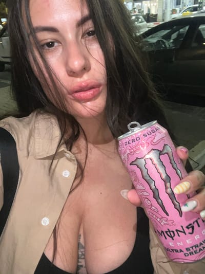 A woman holds a pink can of "Monster Energy Ultra Strawberry Dreams" drink. She has long dark hair and is wearing a beige collared shirt over a black top. Part of her tattoo is visible on her upper chest. The background is blurred and includes what appears to be a building and possibly a vehicle. There is no information in the image to support a location in the city of Kiryat Motzkin, Israel.