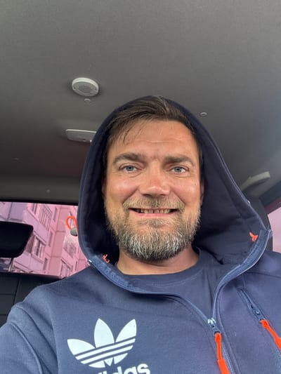 This is a selfie of a middle-aged Caucasian man with a beard and blue eyes, wearing a navy blue hooded jacket with orange zipper pulls. The jacket has an Adidas logo with three stripes and a trefoil on the chest. The man is smiling, showing his teeth. He appears to be inside a vehicle, possibly a bus or RV, as indicated by the ceiling and a circular vent visible on the left side of the image. The background shows a blurred image of buildings with pinkish hues, suggesting it might be daytime with artificial lighting or a filter applied. There is no visible text other than the Adidas logo. The overall emotion conveyed is one of casual happiness. The image is oriented sideways.