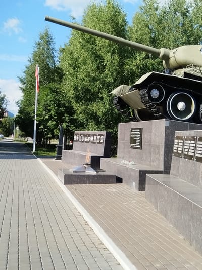 In the city of Roshchinsky, Russia, a monument featuring a military tank and an eternal flame is depicted. The tank, a Soviet-era T-34, is positioned on a raised granite base, its gun barrel extending diagonally across the frame. Adjacent to the tank is a memorial wall with several dark plaques, presumably listing names or significant events. The eternal flame burns brightly in a metallic fixture in front of the wall. The surrounding area is paved with light-colored interlocking bricks, with a clean white curb separating it from a darker paved area. Green trees form a backdrop behind the monument, and a blue sky with scattered white clouds is visible above. A red and white flag is planted on a pole to the left of the monument, behind the paving stones. In the background, a street with a car and a pedestrian walking can be seen.