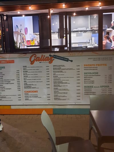 The image shows an outdoor seating area and a takeaway window of a restaurant named "Galiaz," located in Pesaro, Italy. The takeaway window displays a menu with various food and drink items, including pizzas, fried potatoes, beverages, and beers. The menu is presented in Italian. The restaurant's name and a slogan "LA SPECIATA AL MARE" (The sea-speckled) are visible on the menu. The environment appears to be an urban setting, with brick pavement and outdoor furniture. Two individuals are visible through the window of the restaurant, one appearing to be an employee and the other a customer. The lighting suggests it is either dusk or evening.