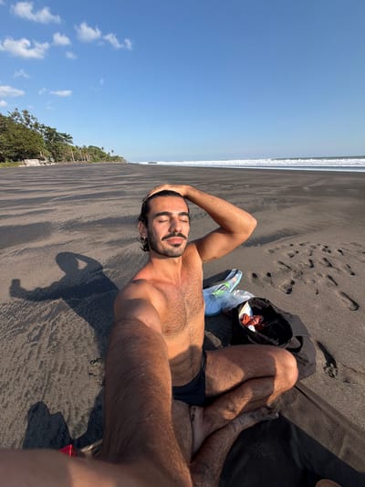 A male individual is depicted in a full-frame selfie, seated cross-legged on a black sand beach in Selemadeg Timur, Indonesia, under a clear blue sky with scattered white cumulus clouds. The man, who appears to be of Mediterranean or Middle Eastern descent, has dark hair, a beard, and a mustache. He is shirtless, wearing dark swim trunks, and has a small silver hoop earring in his left ear. His eyes are closed, and he exhibits a slight smile, with his right hand (viewer's left) resting on his head and his left arm (viewer's right) extended forward holding the camera. A distinct shadow of the man, including his arm and hand holding the camera, is cast on the dark sand to his left. To the man's right, a black open bag contains orange-and-white striped fabric and other items, next to a clear plastic bag holding a white object and a green item. The wide black sand beach extends into the distance, meeting the ocean where white waves break on the horizon. Lush green trees and palm trees line the distant left side of the coastline. The scene is brightly illuminated by direct sunlight.