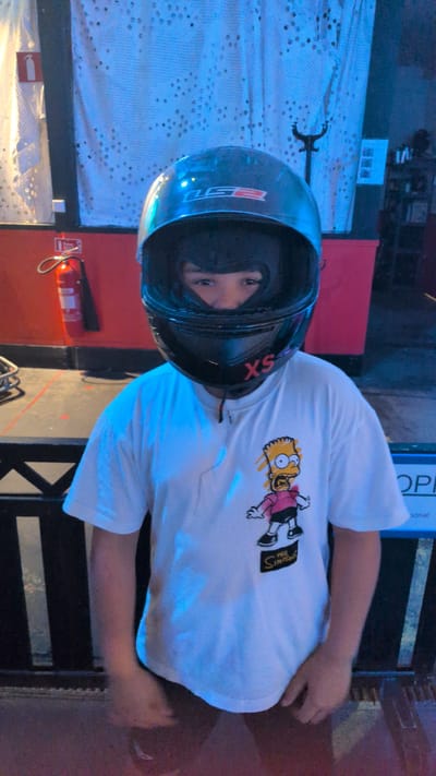A person, likely a child, is wearing a black racing helmet that covers their head and neck. The helmet has a red "XS" sticker on the chin bar. The person is also wearing a white t-shirt with a cartoon image of Bart Simpson from The Simpsons. The background appears to be an indoor venue, possibly an arcade or go-kart track, with a red wall and a white curtain with a patterned design. A red fire extinguisher is visible on the left side of the image. A dark metal fence is in the foreground. The lighting is dim and predominantly blue, suggesting artificial illumination. The person is facing the camera and their eyes are visible through the helmet's visor. The scene does not appear to be located in Östersund, Sweden.