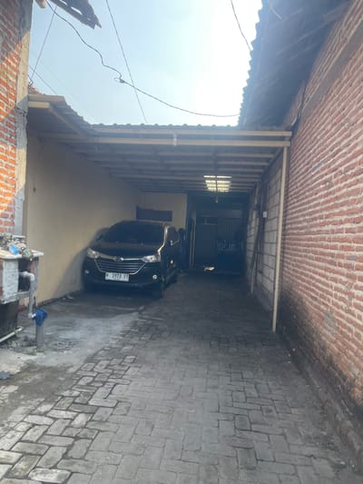 The image depicts a narrow, covered walkway or driveway alongside a brick building in Anggaswangi, Indonesia. A black car is parked within the covered area, facing towards the viewer. The car's license plate reads "W 1973 UT". The ground is paved with interlocking bricks. A portion of a concrete structure with plumbing is visible to the left of the car, with some fabric items placed on it. The walls are made of brick and concrete blocks. Overhead, a corrugated metal roof is supported by beams, with electrical wires strung across the open sky. The scene suggests an outdoor, residential or commercial space, possibly a home with a carport.