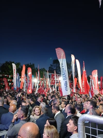A large crowd of people, mostly men, are gathered outdoors in Silivri, Türkiye, at night. The crowd is densely packed, with individuals standing shoulder-to-shoulder. They are gathered in front of a stage or platform where a political rally or event is taking place. Red and white flags with Turkish writing are displayed throughout the scene. The event is illuminated by artificial lighting, and the sky is dark. The crowd appears to be watching and listening to a speaker or performer on the stage.