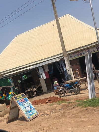The image depicts a street scene in Naraguta Hausawa, Nigeria, featuring a commercial building with a corrugated metal roof. In the foreground, a blue motorcycle is parked on a dirt road. To the left, a three-wheeled auto-rickshaw is visible. An A-frame sign stands prominently in the foreground, displaying images of building materials and the text "SAUKI BUILDING MATERIAL & POS AVAILABLE." The building itself appears to house several small shops, with clothing and other goods displayed outside. Overhead power lines traverse the sky. The overall environment suggests a bustling marketplace or commercial area.