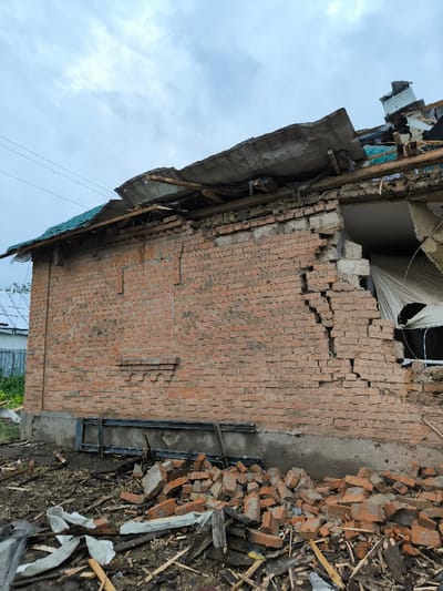 The image depicts a severely damaged single-story structure in Pysarivka, Ukraine, under an overcast sky. The building's exterior is constructed of red brick, with its lower foundation appearing as a darker, possibly concrete or stone, base. The right side of the brick wall exhibits significant structural failure, characterized by a large, irregular void where material has collapsed inward, revealing a dark interior. Jagged, multi-directional cracks propagate extensively through the remaining brickwork in this section. A large, torn white fabric or tarp hangs partially into this opening. The roof is critically compromised, with a substantial portion of grey corrugated roofing material ripped upwards and dislodged, exposing underlying wooden rafters and beams. A section of intact green corrugated roofing is visible on the left side of the structure. The ground immediately in front of the damaged wall is covered with a dense accumulation of debris, predominantly consisting of shattered red bricks, fragmented wooden planks, and torn white material. A dark metal frame or support structure rests against the lower portion of the damaged wall. In the upper left background, electrical or communication lines are visible, spanning across the sky above a light-colored corrugated fence and a partially visible distant structure. No individuals or active processes are present in the scene.
