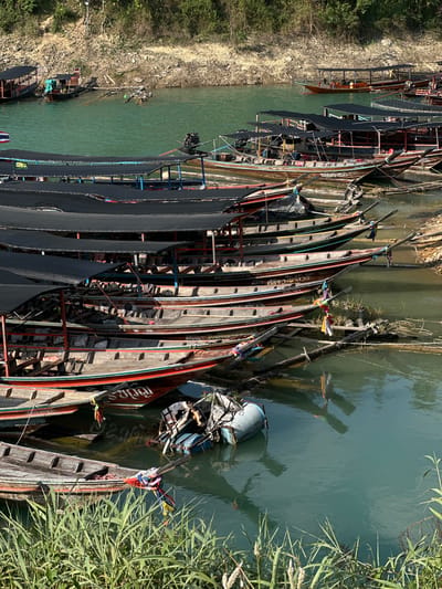 Many boats are docked along the river's edge in Ban Ta Khun, Thailand. The boats are long and narrow, constructed of wood, with some covered in black tarps. One of the boats has the word "phupha" painted in Thai on its side. The water is green-blue. The shoreline is sandy and overgrown with trees and grass. Some tall reeds are growing on the right.