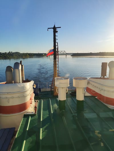 The image depicts the stern view of a ship navigating a wide river or canal, observed from an elevated position on the vessel's deck, with the location identified as Kochino, Russia. The scene is set during early morning or late afternoon, characterized by a clear, light blue sky and a low sun angle from the right, casting long, distinct shadows across the deck.
In the foreground, the ship's green, ribbed metal deck is visible, reflecting some light and showing signs of dampness. To the far left, a section of a dark blue wooden bench or seating is present. Centrally positioned on the deck are two prominent white cylindrical structures, likely vents or air intakes. The left vent is labeled with the Cyrillic text "АККУМУЛЯТОРНАЯ" (AKKUMULYATORNAYA), indicating it's related to the battery or accumulator room. A faint wisp of white vapor or steam is visibly emanating from the top of the right vent.
A tall, dark metal mast with several rungs or steps extends upwards from the central-right portion of the deck. Attached to this mast is a Russian national flag (white, blue, and red horizontal stripes), fluttering gently towards the left. On both the left and right sides, parts of the ship's upper structures are visible, painted white with a distinct red stripe, suggesting these could be the hulls of lifeboats or other superstructure components. Behind the structure on the left, two grey/silver exhaust funnels are visible.
The ship is actively moving, creating a V-shaped wake that trails behind it on the calm, reflective surface of the water. In the mid-distance, a large metal truss bridge, likely a railway bridge, spans the waterway. Further beyond, a second, simpler bridge or overpass can be faintly discerned. The riverbanks are lined with lush green foliage, interspersed with some distant buildings, utility poles, and what appears to be a cleared area or road on the left bank. A light mist or haze hangs low over the water near the distant right bank. No individuals are visible in the frame.