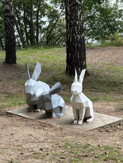 The image depicts a park scene in Zarechny, Russia, featuring two geometric sculptures of rabbits. The sculptures are made of polygonal shapes, with one rabbit being white and the other a dull gray. They are positioned on a concrete slab in a wooded area. The ground around the slab is covered with dirt, scattered leaves, and some patches of grass. In the background, several trees with dark bark are visible, along with foliage that suggests a forest or park setting. The lighting indicates it is daytime. There are no people or animals present in the scene.