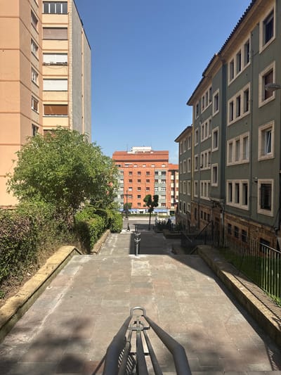 The image shows a view looking down a wide stone staircase in Oviedo, Spain. The staircase is flanked by buildings on both sides. On the left is a tall, peach-colored building with many windows, some of which are covered by blinds. Adjacent to this building is a lush green tree and dense hedges. On the right is a lower, green-painted building with many windows framed in a light color. A black metal fence runs along the edge of the sidewalk next to this building. The staircase itself is made of large, square stone tiles with a reddish-brown hue. Metal railings are present at the bottom of the stairs and on either side, though the focus of the foreground is on a section of railing at the bottom. In the distance, at the end of the staircase and beyond the road, is a multi-story brick building with a red tiled roof. The sky is a clear, bright blue. A single person is visible walking down the street in the distance, and shadows from unseen objects cast dark shapes on the staircase. The scene appears to be a public space within a residential area, likely a street or plaza connecting different levels of the city.