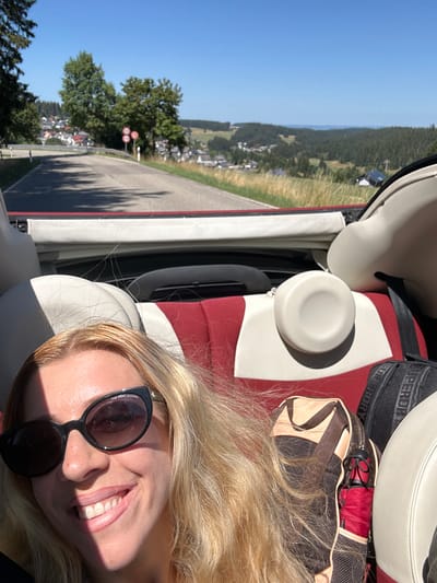 A blonde woman with sunglasses smiles at the camera while sitting in the passenger seat of a convertible. She is in the city of Schönwald im Schwarzwald, Germany, on a sunny day. The car is on a road that winds through a scenic landscape of rolling hills, forests, and houses. The woman has long, blonde hair, and she is wearing dark sunglasses. The car's interior is visible, with red and white seats. There are bags in the back seat. The background shows a clear blue sky and a lush green countryside. The road has a red stripe running along its edge.