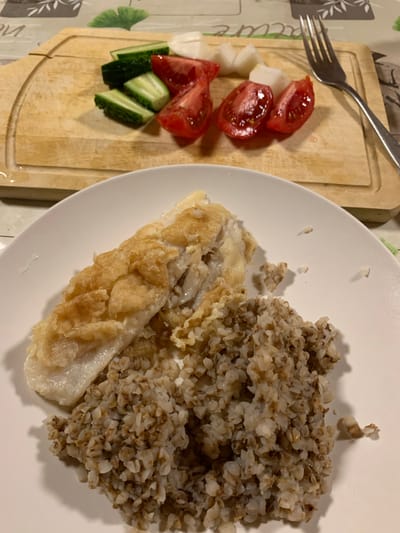 The image displays a homemade meal arranged on a table indoors. On the right, a white round plate holds a serving of cooked buckwheat, characterized by its granular, light brown and beige appearance, and a piece of fried or pan-seared fish, which appears to be coated in a light-colored batter or breading.

Adjacent to the plate, on a light-colored wooden cutting board, are components for a side salad. These include several wedges of fresh red tomatoes, diagonally sliced green cucumbers, and some white cubed items, possibly cheese, radish, or onion. A silver fork is placed on the cutting board, partially overlapping the white cubes.

The table is covered with a patterned tablecloth featuring faint, partially legible text on the left, but the full words cannot be deciphered. The lighting suggests an indoor setting, likely under artificial light, with soft shadows visible on the plate and cutting board. The scene depicts a simple, prepared meal, ready for consumption. No individuals are visible in the image.
