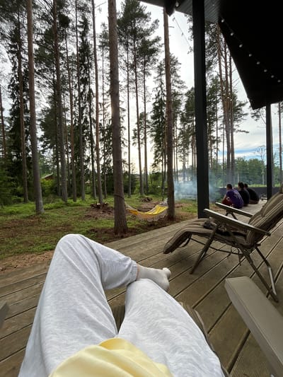 The image depicts a scenic view from a wooden deck in Shepelyovo, Russia. In the foreground, a person's legs clad in light grey sweatpants and white socks are visible, resting on the deck. A yellow garment is also partially visible. The deck overlooks a lush green forest with tall, slender pine trees. A yellow hammock is strung between two trees in the middle ground. Further in the background, a plume of smoke rises from a fire pit, suggesting a gathering or cooking activity. Several individuals are seated on chairs on the deck, observing the scene. The overall ambiance is one of relaxation and natural beauty.