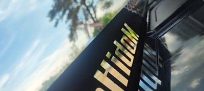 The image is an abstract, angled shot of a black object with metallic lettering, viewed through a reflective surface. The lettering appears to spell out "Hoidok". The reflective surface shows a blurred background of sky with clouds and trees, suggesting it is either a window or a glossy surface outdoors. A portion of a black plastic object, possibly a car interior component or electronic device casing, is visible on the right side. The image is taken from a low angle, looking upwards. The overall impression is of a close-up, somewhat distorted perspective, focusing on the texture and sheen of the materials. There is no discernible location in the city of Ia Hru, Vietnam, or any people or specific actions depicted.