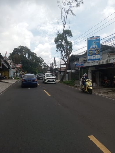 A street scene captured during daylight. In the foreground, an asphalt road stretches forward, marked by yellow lane lines. A light yellow scooter with two people aboard travels along the right side of the road. Behind the scooter are several cars. On the left, a blue sedan is followed by other vehicles. On the right side of the road, storefronts are visible, including one with a sign that reads "INDO LAUNDRY" and another with "75 CUCI KERING MENIT SELESAI". Trees and utility poles line the street. The sky is overcast.
