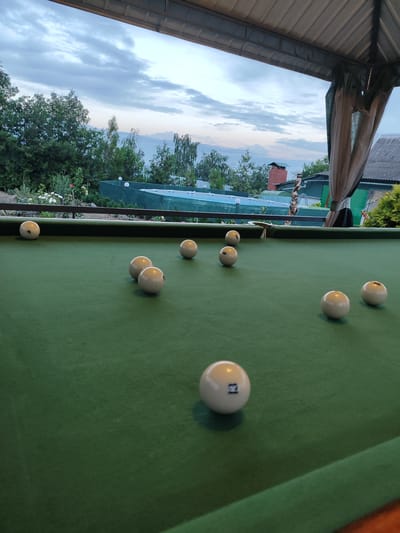 This image depicts an outdoor billiard table under a canopy, likely in Krasnokutsk, Ukraine, given the reference. The table is covered in green felt, with several billiard balls scattered across its surface. The balls are white or off-white, and some have visible numbers. In the background, there is lush green foliage, a fence, and residential structures. The sky is overcast with dramatic clouds, suggesting it might be late afternoon or early evening. The scene appears to be a relaxed, possibly recreational setting.