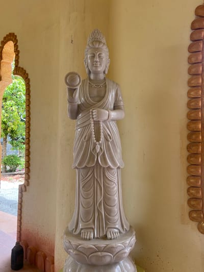 A full-body standing statue, crafted from light-colored stone, likely marble, is centrally positioned against a smooth, light yellow wall in the city of Đăk Mar, Vietnam. The statue depicts a serene Buddhist deity, possibly Guanyin, adorned with an ornate crown, flowing robes, a beaded necklace, and bracelets. Its raised right hand holds a disc prominently featuring the Yin-Yang symbol. The left hand holds a string of white prayer beads (mala) with a tassel, draped downwards. The statue rests on an elaborate lotus-shaped pedestal. To the immediate left, an arched opening framed by a reddish-brown textured border provides a view of an outdoor environment with lush green foliage and a paved ground. A dark, cylindrical object is situated on the ground near the base of this archway. A similar reddish-brown textured architectural element is partially visible along the far right edge of the frame.