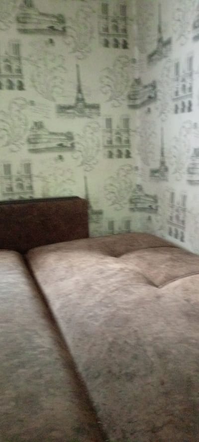 The image shows a close-up view of a couch with a brown backrest and a textured, light brown seating area. The background features wallpaper with a pattern of architectural sketches, including what appears to be the Eiffel Tower and other Parisian landmarks. The lighting is dim and appears to be from an interior light source. There is no indication of the city of Terpinnia, Ukraine, or any people present in the scene. The focus is on the couch and the wallpaper.