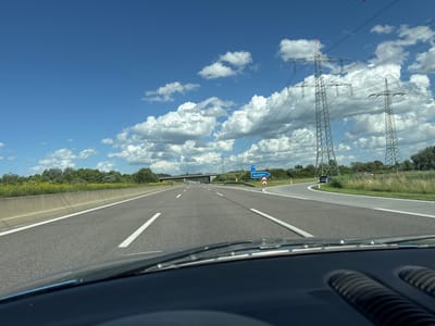 The image captures a forward view from inside a vehicle on a highway, likely in Friedberg, Germany, given the presence of a blue directional sign indicating "Ausfahrt" (Exit) and "U 416" (an indication of a state road). The highway has multiple lanes, marked with white lines. On the right side of the highway, a blue sign points to an exit. Large pylons supporting power lines are visible on the right side of the road, stretching towards the horizon under a bright blue sky dotted with white, fluffy clouds. Lush green trees and vegetation line the sides of the highway, with a concrete barrier separating the lanes. The dashboard of the vehicle is partially visible in the foreground. The weather appears to be clear and sunny.