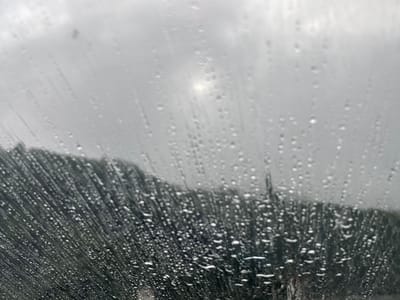 A close-up, low-angle shot shows raindrops on a windshield, obscuring a blurry background of a overcast sky and a dark, tree-covered hillside. A bright spot of light, possibly the sun peeking through the clouds, is visible in the upper center of the frame. The lower portion of the image is dominated by the textured pattern of water droplets and streaks running down the glass, creating a sense of depth and atmosphere. The overall impression is one of being inside a vehicle during a rain shower in a natural setting, possibly on a mountain road or in a rural area near Tsakva, Georgia. There are no visible people or specific identifiable landmarks, and the focus is entirely on the visual effect of the rain on the windshield and the obscured landscape beyond.