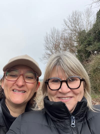 A photograph taken outdoors in Saint-Cyprien, France, depicts two adult females positioned closely together, smiling directly at the camera. The individual on the left has dark hair, wears a beige baseball cap, and large brown-framed eyeglasses; her mouth is open in a smile revealing her teeth. The individual on the right has blonde and grey hair with bangs, wears dark, possibly tortoise-shell framed eyeglasses, and smiles with a closed mouth. She is dressed in a black puffer jacket with a prominent silver zipper. The background consists of an overcast grey sky above a rural or semi-rural landscape featuring bare deciduous trees, some evergreen foliage on the right, and an expanse of dry, light-brown vegetation, possibly a field, behind the subjects.
