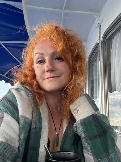 A woman with vibrant red, curly hair is taking a selfie outdoors. She is wearing a green and white plaid jacket and a necklace with a green pendant. The background shows a blue awning and part of a building with windows. The image appears to be taken on a boat, possibly in Zuevo, Russia, given the presence of a blue awning commonly found on Russian boats.