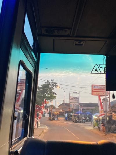 The image provides a forensic view of a street scene in Parungserab, Indonesia, captured from the interior of a vehicle, likely a bus, looking through its front windshield. The sky overhead is a clear blue with a lighter, hazy horizon and scattered high clouds, suggesting either early morning or late afternoon.

In the immediate foreground, the dark interior of the vehicle is visible, including what appears to be a dashboard or seat top. The vehicle's ceiling is present, featuring overhead panels and ventilation grilles. The upper section of the windshield exhibits a prominent blue-green tint. A partial "ATM" logo is visible, likely as an internal decal or reflection, on the upper right portion of the windshield.

Beyond the windshield, a two-lane asphalt road extends into the distance. On the left side of the road, a sidewalk or shoulder is lined with numerous red and white Indonesian flags, suggesting a national observation or event. A large green tree stands further along the roadside. An illuminated streetlamp is visible, and beneath it, several temporary market stalls or vendor booths, some covered by blue tarpaulins, are set up. Multiple individuals are present near these stalls, and several motorcycles are parked on the roadside. A large, legible billboard in the distance on the left displays "SMP UNGGULAN MA'ARIF CENDEKIA."

On the right side of the road, more red and white flags are displayed. A prominent "Alfamart" convenience store sign, featuring red lettering on a white background, is visible above a cluster of buildings and additional temporary market stalls. More individuals and parked motorcycles are observed on this side of the street. Structures with dark roofs line both sides of the thoroughfare. The scene depicts an active urban or semi-urban street environment with commercial activity.