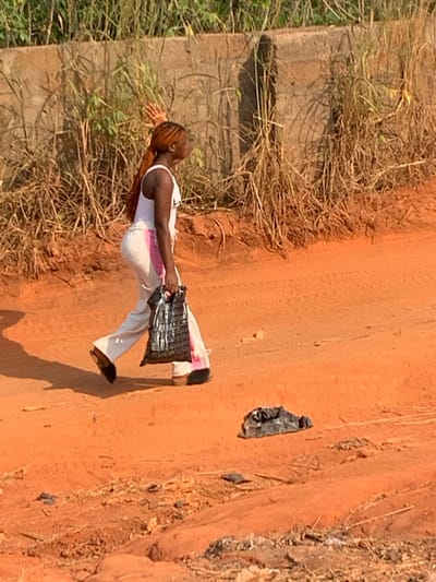The scene is an unpaved outdoor environment in Nando-Ikem, Nigeria, characterized by reddish-brown dirt. A single individual, a female with long, reddish-brown hair styled in a ponytail, is depicted walking from left to right across the frame. She wears a white sleeveless top and light-colored pants featuring a vertical pink stripe along the side. Her left hand is raised, palm partially visible, above shoulder level. Her right hand holds a crinkled black plastic bag. Her head is turned slightly to the left, and her mouth is open. In the background, a weathered concrete wall rises, partially obscured by dry, tall grasses and weeds. Another crinkled black plastic bag lies on the dirt ground to the right of the walking individual. Faint tire tracks are visible in the dirt. The lighting indicates daytime conditions.