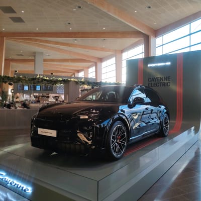 A black Porsche Cayenne Electric SUV is prominently displayed on a tiered, dark gray platform in a spacious, modern indoor environment at Stockholm-Arlanda, Sweden. The vehicle is positioned facing left at a slight angle towards the viewer, with its front left quarter visible. A small white sign bearing the word "Cayenne" in script is attached to the front lower grille. Below the car, on the front edge of the display stand, the word "Cayenne" is illuminated in blue neon-like script.
Behind the vehicle, a large black wall serves as a backdrop, featuring the illuminated white text "PORSCHE" at the top, and below it, "NYA CAYENNE ELECTRIC." Two vertical red stripes with a glowing effect flank the text. The car's polished black surface reflects the surrounding ceiling, windows, and display elements.
The architectural setting features a high ceiling constructed from light-colored square acoustic panels with a subtle grid pattern, interspersed with numerous recessed circular lights and air vents. Robust, light-colored wooden beams form an intricate, angular support structure across the ceiling and extend downwards as columns. Large windows along the right side of the space admit abundant natural light, reflecting off the shiny surfaces.
In the background, to the left of the car display, a commercial food and beverage establishment is visible. A prominent sign with a mountain-like logo reads "NOR KITCHEN." Below the sign, a long counter area is equipped with menu boards, various bottles, and glassware. A decorative green garland with small lights hangs above the counter. Several individuals are present at this counter: two men are seated, one standing, and a staff member in an apron is positioned behind the counter, engaged in service. The floor throughout the area consists of dark, rectangular tiles.