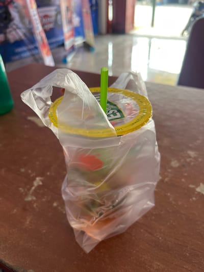 A beverage in a plastic bag sits on a wooden surface. The beverage is in a clear plastic cup with a yellow lid. A green straw is visible sticking out of the cup. The cup is partially obscured by the plastic bag, which also contains what appears to be ice and possibly fruit. The lid of the cup has text printed on it. Some visible text on the lid is "Fresh Brew" and "Tanpa Pengawet". The background shows shelves with various items, suggesting a retail or convenience store setting. The lighting suggests it is daytime, possibly indoors with bright artificial light or near a window. The overall scene is casual and everyday.