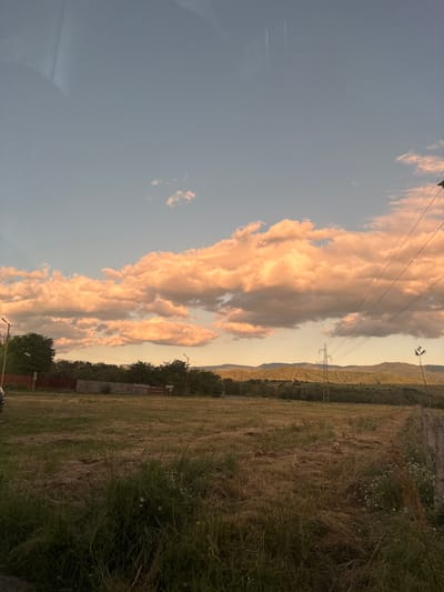 The image captures a rural landscape near Zlatitsa, Bulgaria, bathed in the warm light of what appears to be sunset. The sky is a blend of pale blue and soft peach hues, dominated by large, fluffy clouds illuminated from below, giving them an orange glow. In the foreground, a field of dry, cut grass stretches across the scene, interspersed with patches of green vegetation and a few small white flowers.

Beyond the field, a line of trees forms a dark green border, with a rust-colored fence visible in the middle ground. Several utility poles with power lines are scattered throughout the landscape, some reaching towards the illuminated clouds. In the distance, rolling hills covered in trees create a layered backdrop, with the faint outlines of mountains visible on the horizon. The overall impression is one of natural beauty and tranquility in a semi-rural setting.