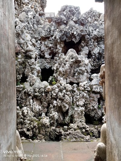 The image shows a stone wall made of rough, gray, and white rocks, with some small, dark crevices and green moss patches. The wall is viewed through a narrow opening, framed by two vertical concrete walls. The ground in front of the wall is made of reddish-brown tiles, with a white line running across it. The location, based on the coordinates provided, is likely somewhere in the city of Cirebon, Indonesia. There are no people or animals in the image.