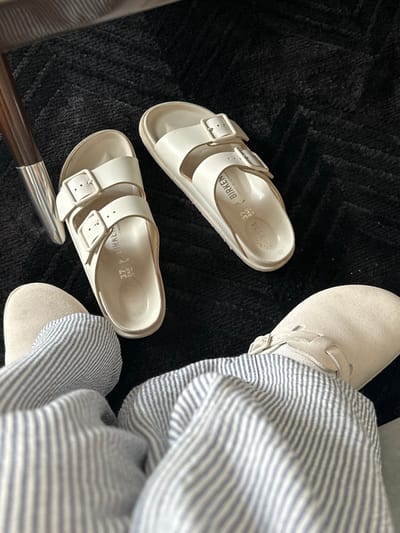 The image depicts an indoor scene in Vandœuvres, Switzerland. A low-angle view focuses on a person's lower legs and feet, clad in light beige, closed-toe clogs with a buckle, and light-colored, vertically striped trousers. Directly in front of the person's feet, a pair of white Birkenstock Arizona sandals, marked size "37 240" and "BIRKEN MADE IN GERMAN", rests on a dark, possibly black, textured carpet with a subtle chevron or geometric pattern. A portion of a wooden furniture leg with a silver-toned metallic foot is visible on the left side of the frame.