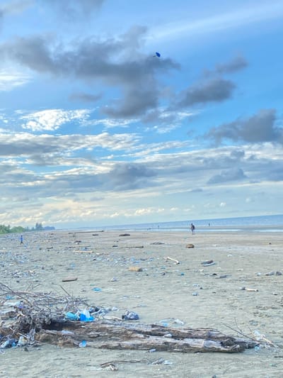The image displays a wide-angle view of a heavily littered sandy beach in Kualaidi, Indonesia, under a partly cloudy sky. The sky occupies the upper portion, featuring large grey and white cumulus clouds interspersed with patches of clear light blue. A small, bright blue kite is visible flying high in the upper right quadrant of the sky.
The foreground is dominated by dark, damp sand strewn with significant natural debris, including large, weathered driftwood logs and piles of tangled branches. Embedded within and around this natural material is a dense accumulation of artificial refuse, primarily plastic bottles and bags, many appearing light blue, white, or dark.
Moving towards the midground, the expansive sandy beach stretches into the distance, maintaining a consistent scattering of smaller driftwood pieces and various plastic items. Several shallow pools or wet sand areas are visible closer to the ocean.
Multiple human figures are present in the midground and background. On the far left, near a distant treeline, a small figure, likely a child in light clothing, walks leftwards away from the camera. Closer to the center, another individual walks towards the ocean, carrying a long, thin pole or stick vertically. Farther out along the waterline, several other extremely distant, indistinct figures are visible.
The background consists of the calm, light blue-grey ocean meeting the horizon. A faint, low-lying landmass or distant shoreline is discernible on the far left horizon, bordered by a strip of green vegetation. The scene is illuminated by diffuse daylight, with no strong shadows.