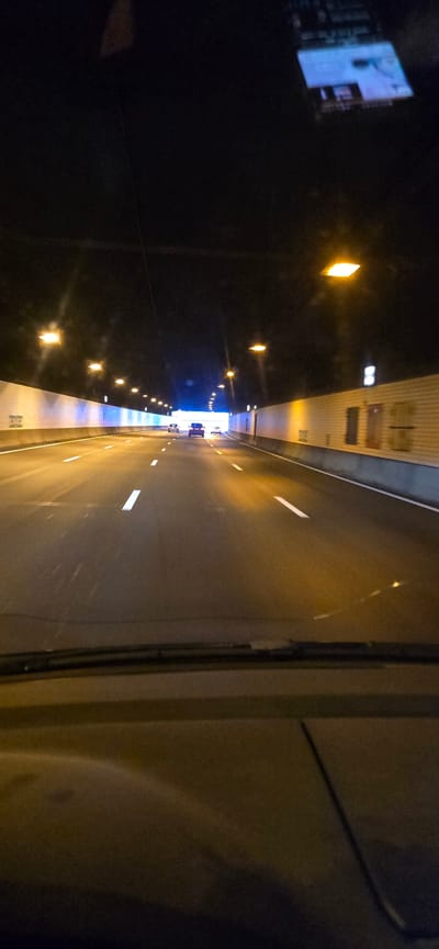 The image shows a view from the interior of a vehicle driving through a tunnel at night. The tunnel has a dark ceiling with several lights illuminating the road ahead. The road is asphalt, marked with white lane dividers. On the sides of the tunnel are white walls with some textured elements. Several cars are visible on the road, traveling in the same direction as the viewer's vehicle. One of the cars is a dark SUV, and another appears to be a white sedan. The image is likely taken on the A9 highway near Badhoevedorp, Netherlands, as this is a major route that passes through tunnels. The reflection on the windshield shows some electronic display, possibly from a dashboard or navigation device. The lighting in the tunnel is a mix of warm yellow and cooler white, creating a somewhat surreal atmosphere.