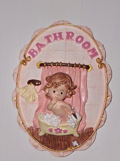 This is a decorative plaque that says "BATHROOM" in pink lettering. The plaque features a pink background with a textured pattern and a gold border. In the center, a small figurine of a baby is sitting in a bathtub filled with bubbles. The baby is holding a brush. A pink curtain hangs behind the bathtub. A cream-colored towel is draped over a hook to the left of the curtain. The plaque is oval-shaped with a scalloped edge. The plaque is mounted on a white wall. The location in Åsgårdstrand, Norway is not visible in the image.