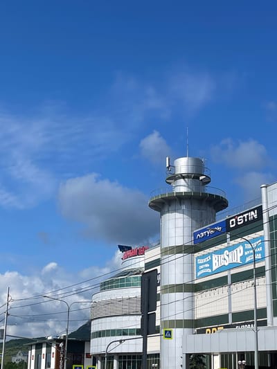 This is a photograph of a modern shopping center in Mineralnye Vody, Russia. The building has a distinctive cylindrical tower structure with a metal exterior and a railing around its upper section. The tower is topped with an antenna. Adjacent to the tower is the main facade of the shopping center, which is also clad in metal and glass. Several prominent signs are visible on the building. One sign reads "O'STIN" in large white letters. Another sign, in blue and white, says "BigShop" with smaller text below that includes "ОДЕЖДА" (clothing) and "ОБУВЬ" (shoes). Above the "BigShop" sign, another sign reads "ЛЕТУАЛЬ" (Letual), a cosmetics retailer. A red sign with Cyrillic lettering is visible on the curved upper section of the building. The sky above is blue with scattered white clouds. Power lines and streetlights are present in the foreground, indicating an urban environment. A mountain range is visible in the background, suggesting the city's geographical setting. There are no people or vehicles clearly visible in the frame. The lighting suggests it is daytime.