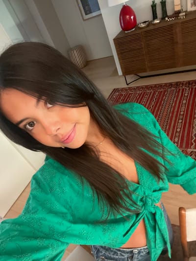 The image captures a young adult female in a selfie format, positioned in the foreground of an indoor residential setting. The specific location within Lordelo do Ouro e Massarelos, Portugal, cannot be determined from the visual information provided.
The subject has long, straight, dark brown hair, dark eyes, and a light-to-medium skin tone, exhibiting a subtle smile and direct eye contact with the camera. She is dressed in a textured, emerald green, long-sleeved, wrap-style crop top with a tie-front closure, revealing a portion of her midriff. Blue denim jeans are partially visible. A delicate gold-colored necklace with a small pendant is worn.
The background reveals a contemporary room featuring light-colored walls and light-toned wooden flooring. A red rectangular rug with intricate geometric patterns covers part of the floor. To the right, a wooden sideboard with slatted doors and black metal legs supports a red oval table lamp with a white shade, two tall dark green decorative vases, a silver-colored bowl, and a small checkered object. The upper left background shows a framed abstract artwork on the wall and a vertically striped cylindrical stool or decorative item. A section of a light-colored wooden chair with a white seat is visible in the bottom right corner. The environment is well-lit by ambient light.
