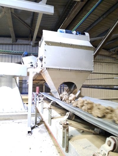 The image shows a conveyor belt system in a large industrial facility, likely a factory or processing plant. Located in Pangasia, Bangladesh (latitude: 22.693, longitude: 90.601), the system comprises a metal hopper with a motor attached, connected to a metal conveyor belt. The belt is transporting a mass of loose, light-brown material, possibly sand or gravel, which is being discharged from the hopper. The environment is characterized by the presence of metal beams, a concrete floor, and a partially visible corrugated metal wall. There are no people present in the image. The system appears to be in operation, as the material is being transported along the belt. The scene is devoid of any human activity.